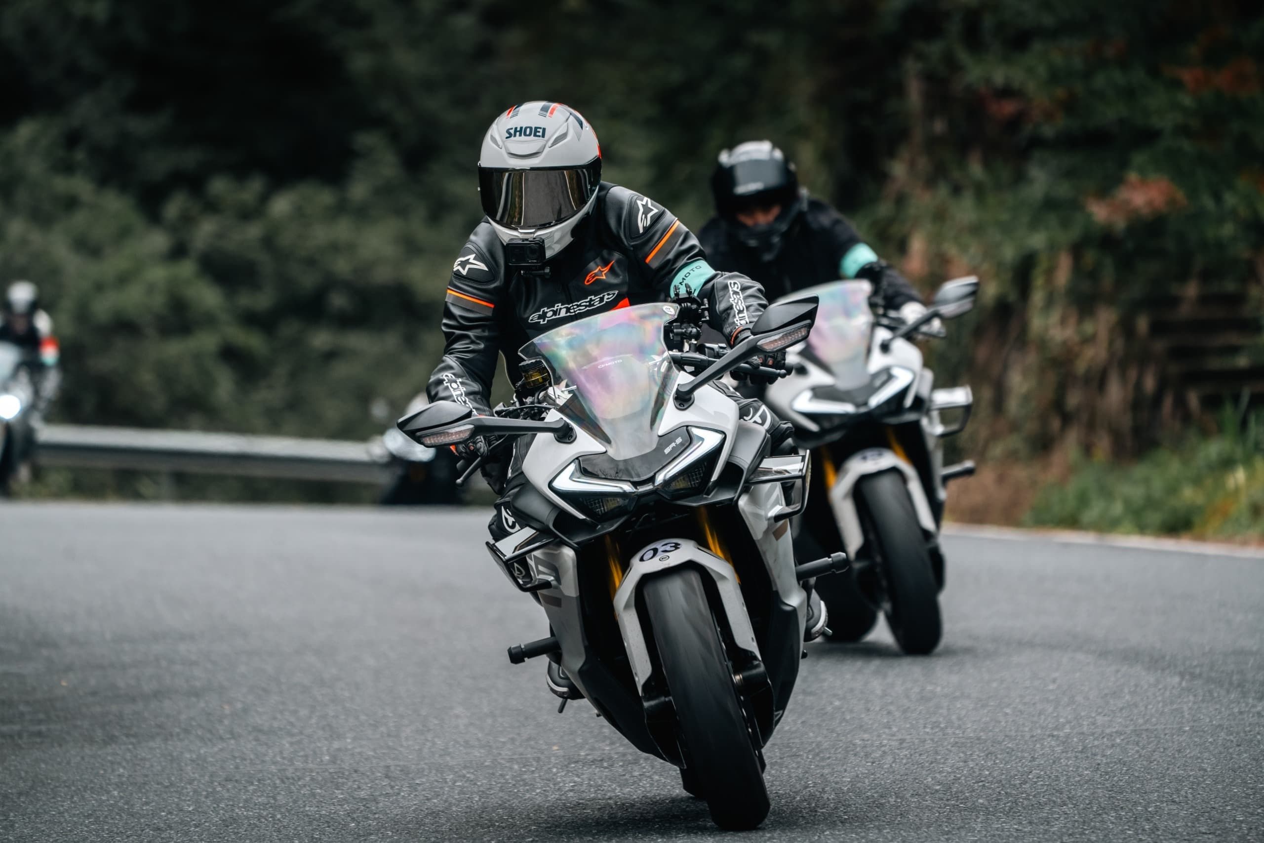 Two motorcyclists in full gear lean into a turn on a winding paved road.