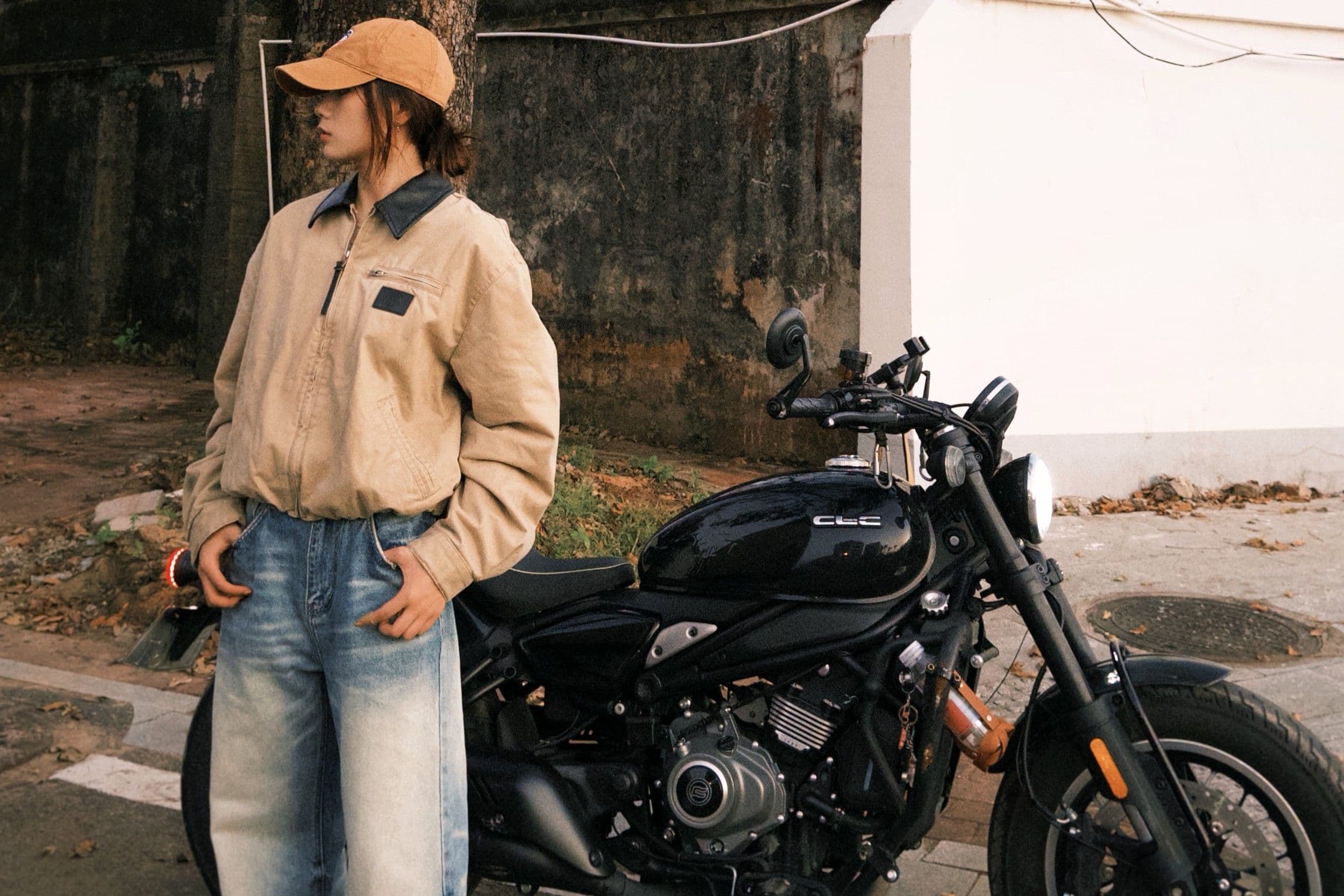 Woman in a cap and light jacket stands beside a black motorcycle outdoors.