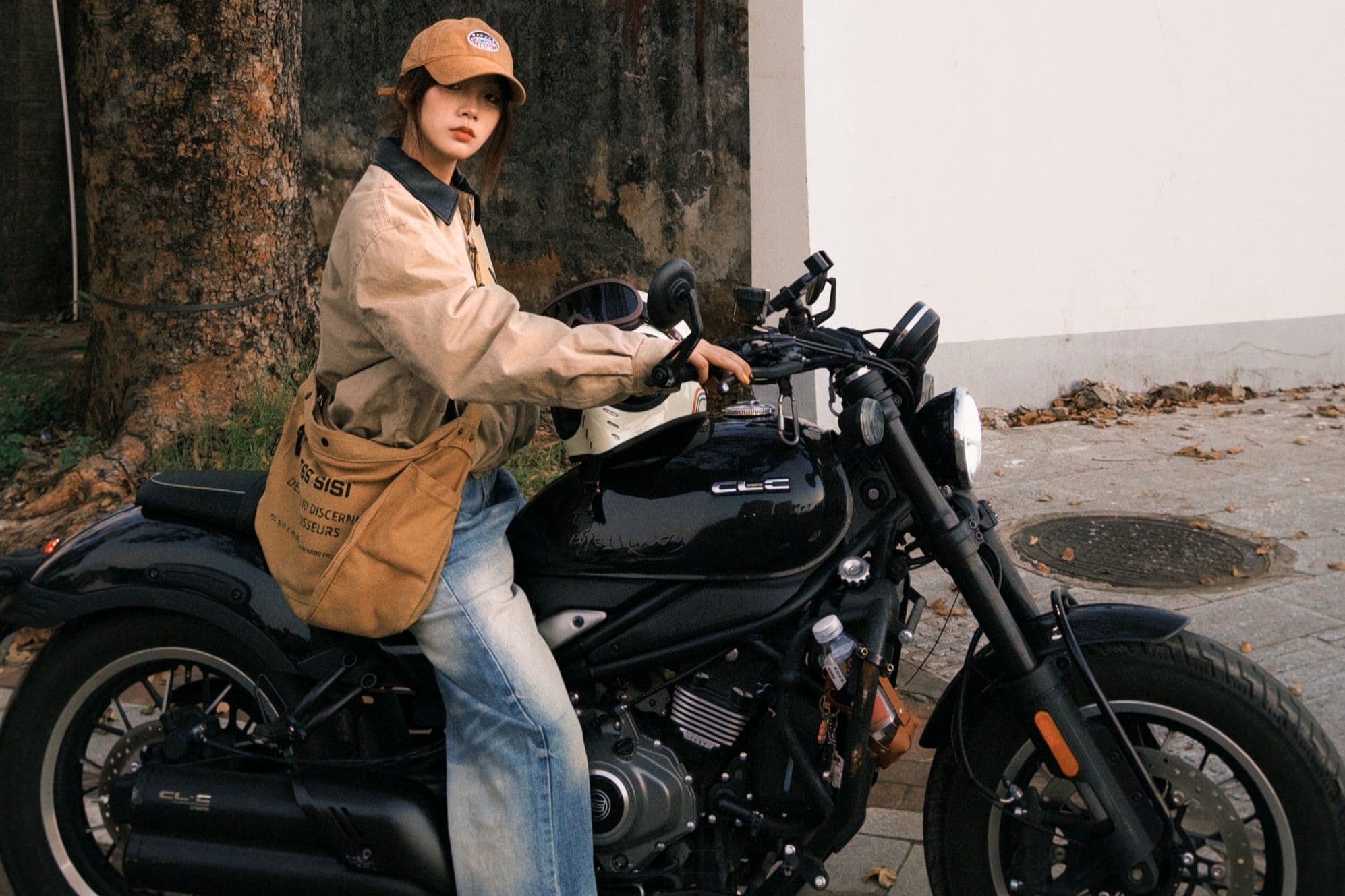 Woman in cap and jacket sits confidently on a black motorcycle outdoors.