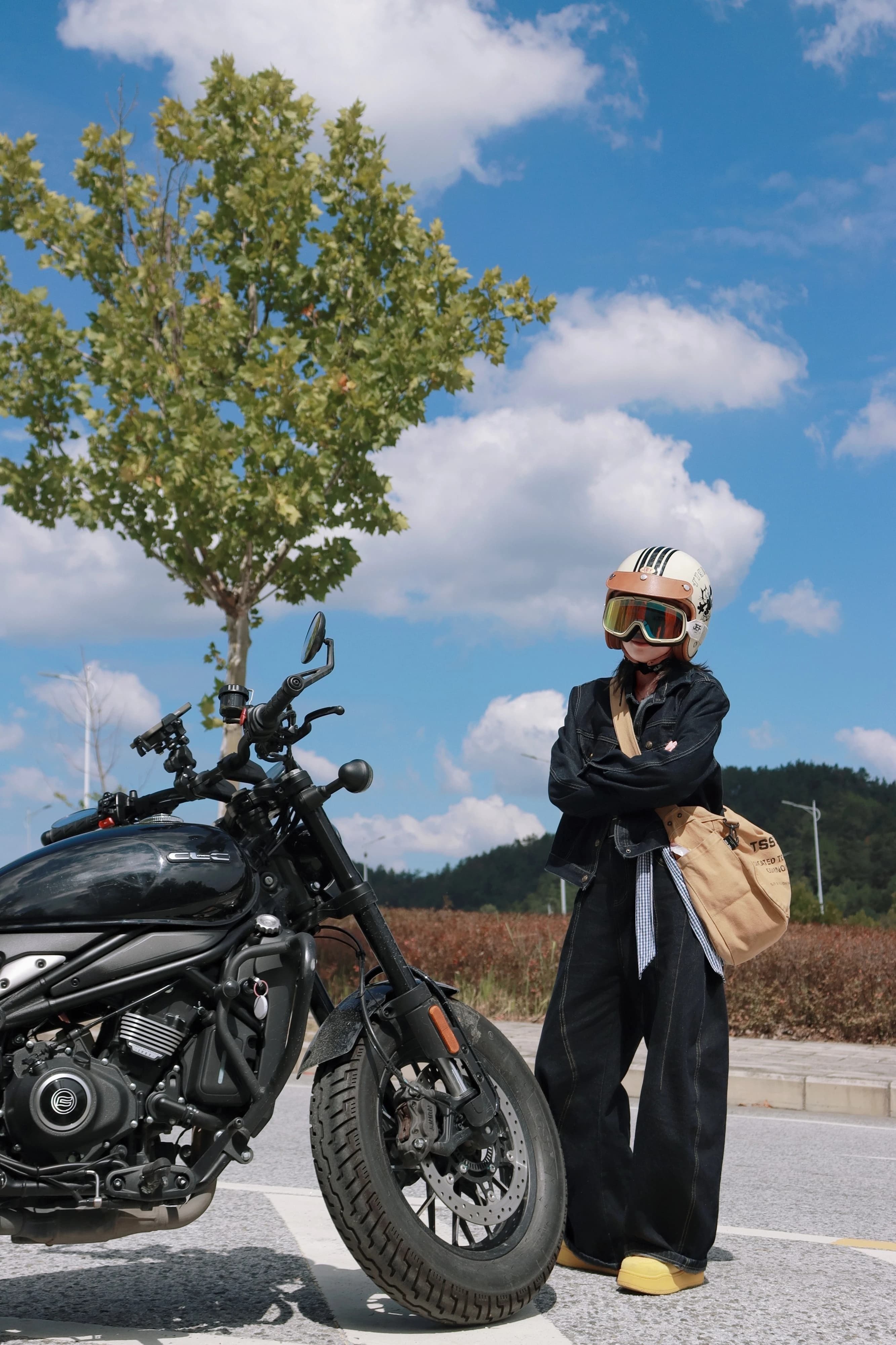 Person in black jumpsuit stands by black motorcycle under tree on sunny day.