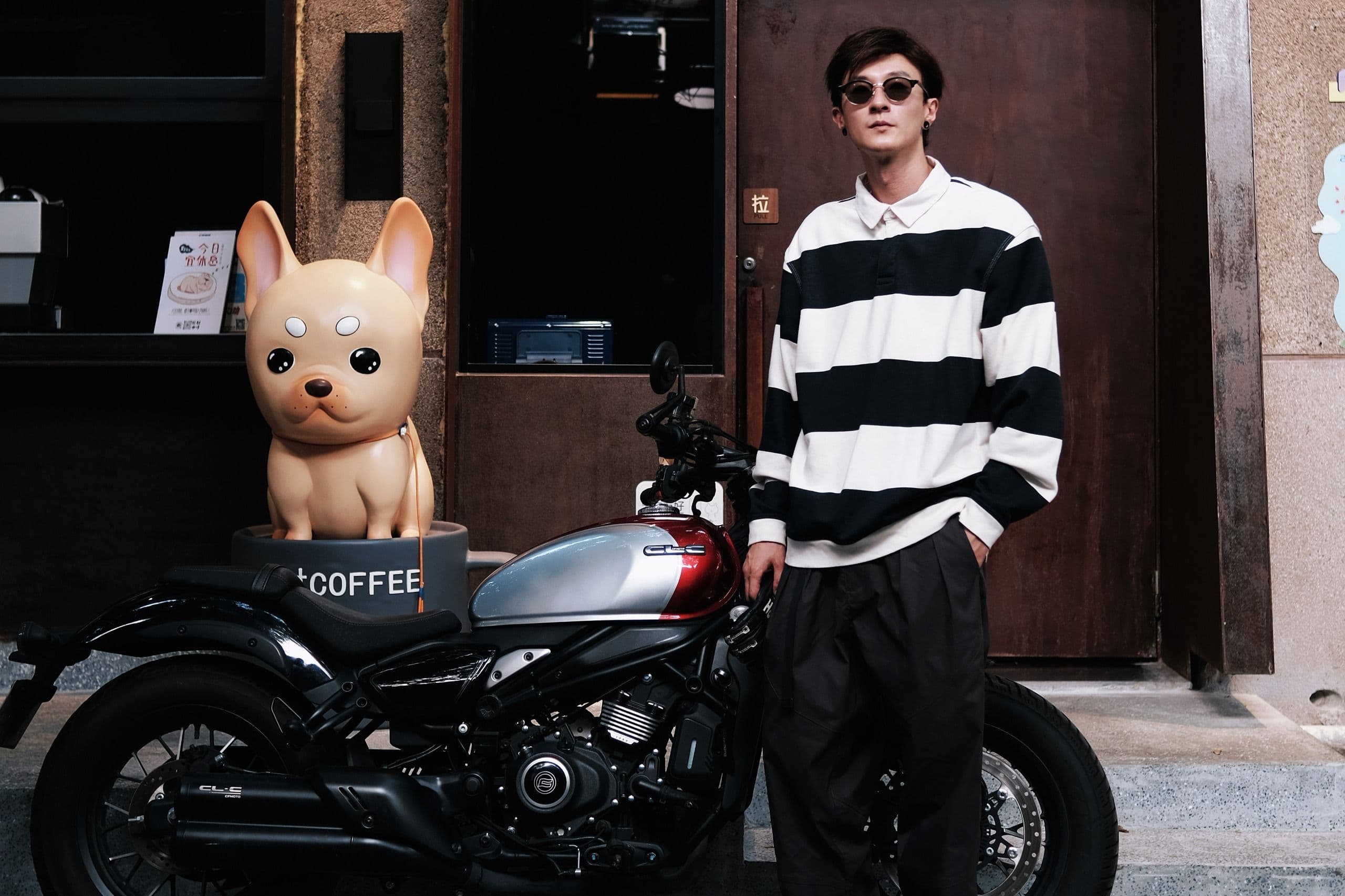 Man in striped shirt and sunglasses stands beside a black motorcycle and dog statue.