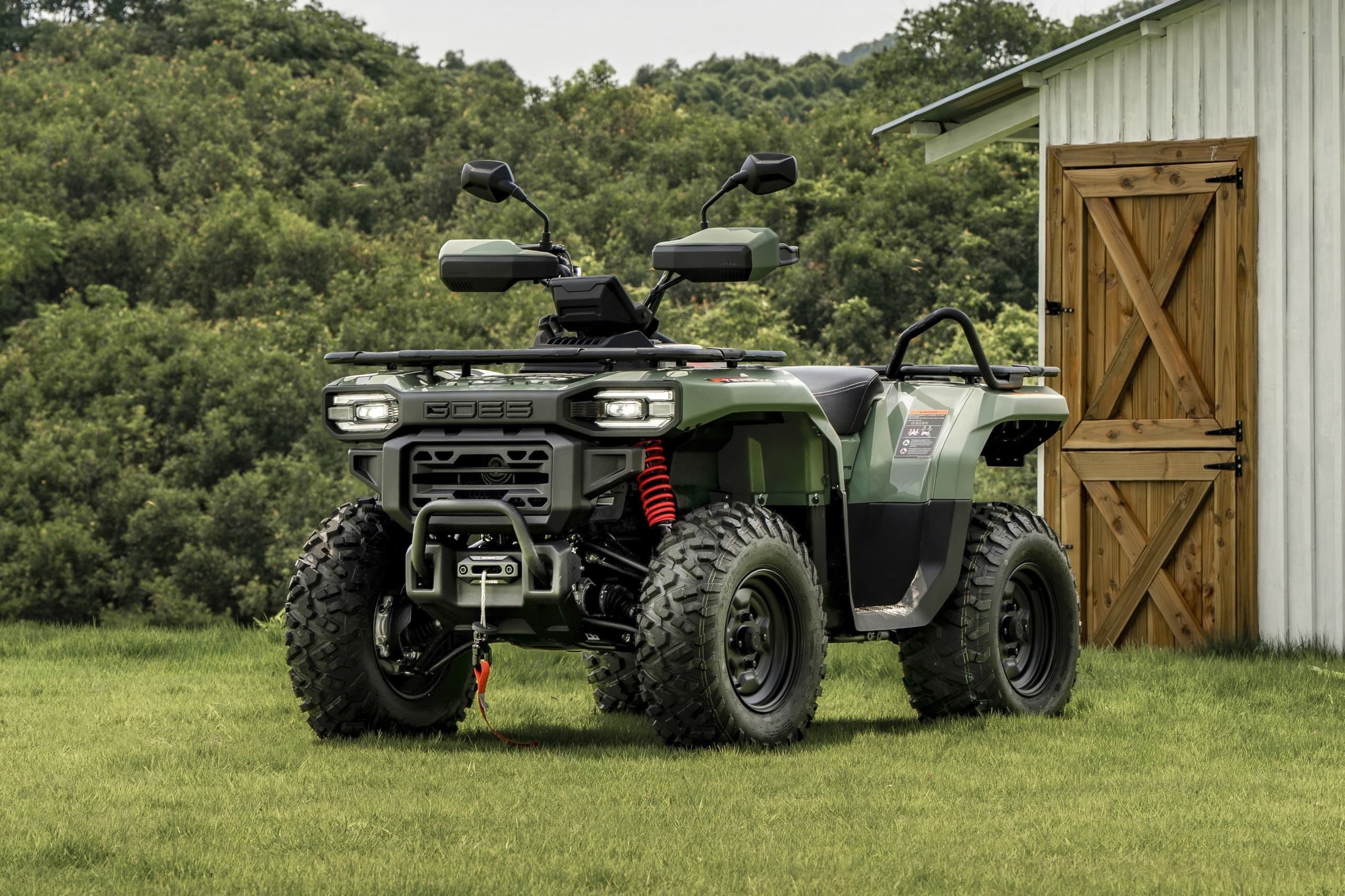 Green camouflage ATV with black tires parked in a lush green field near a barn.