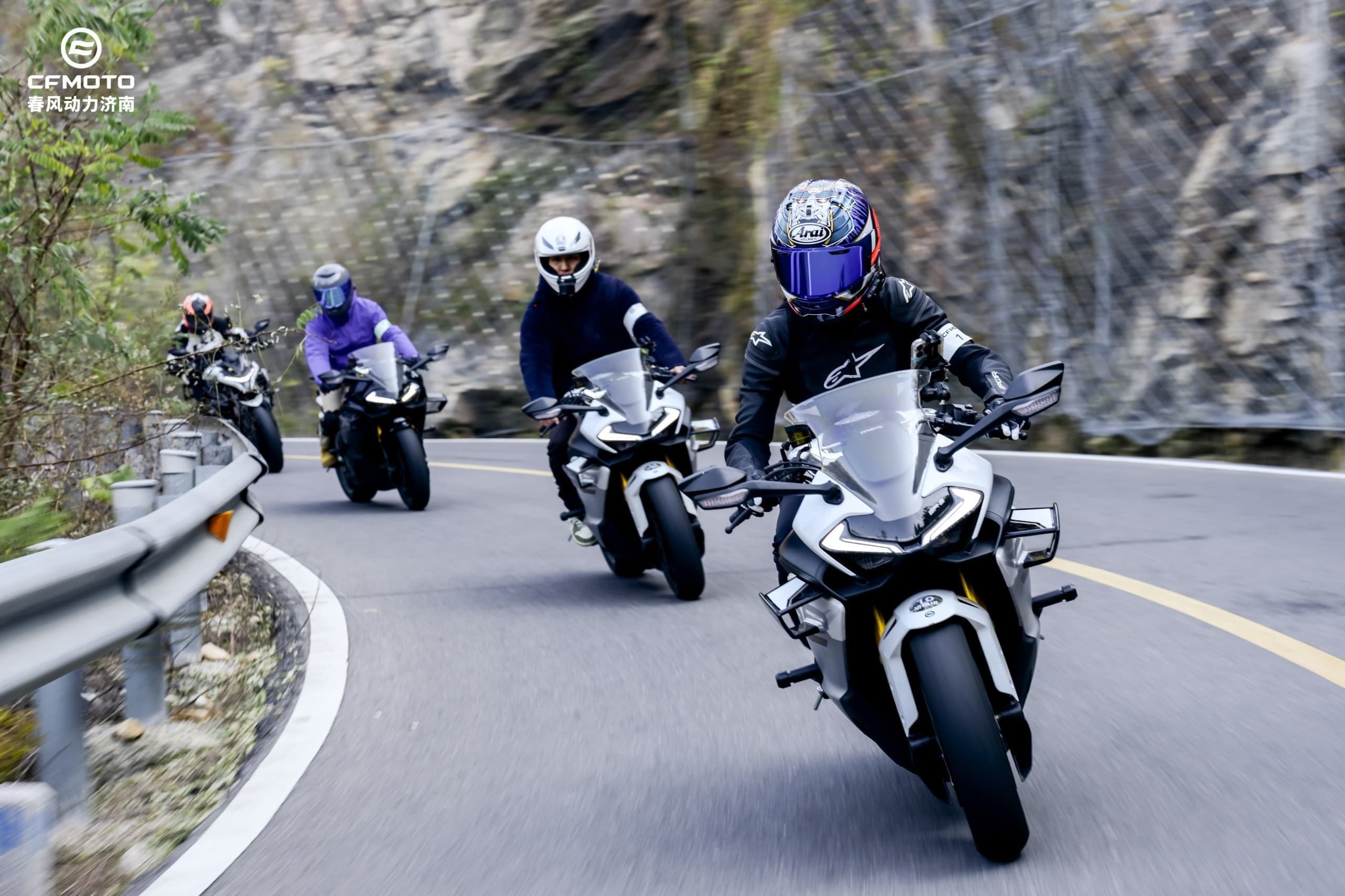 Four motorcyclists ride on a winding mountain road, with the lead rider leaning into a turn.