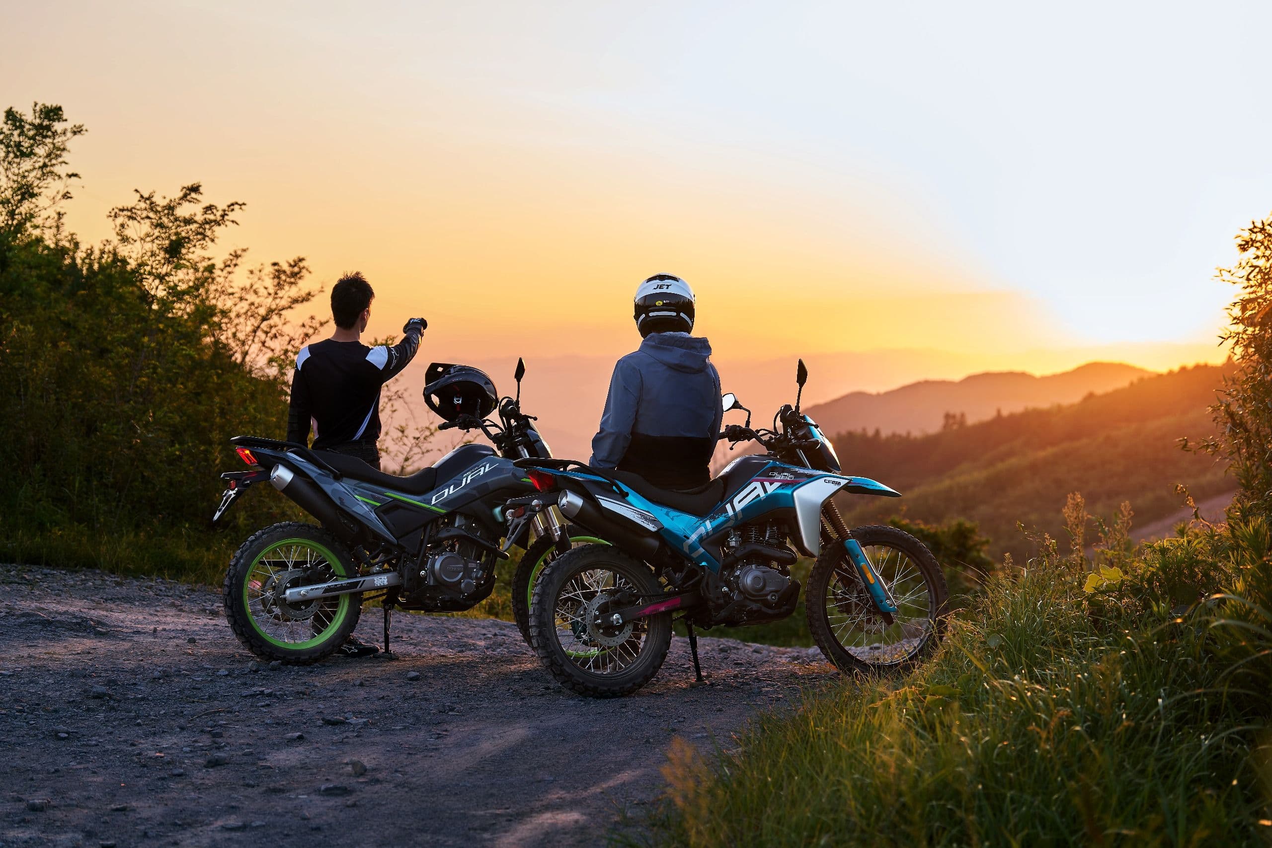 Two riders stand with their dirt bikes on a hill, observing a beautiful sunset over mountains.