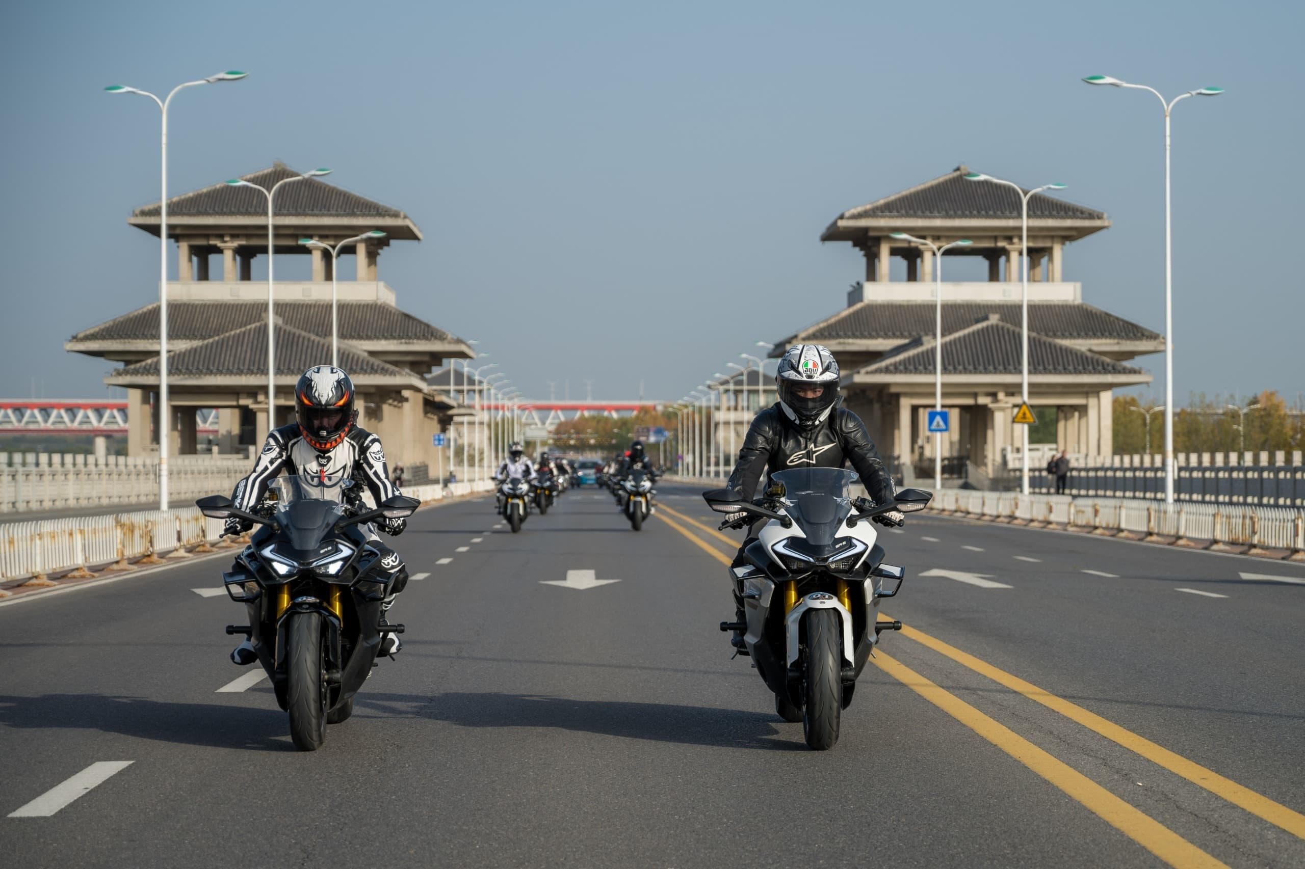 Two motorcyclists approach ornate traditional Chinese gate structures on a wide road.