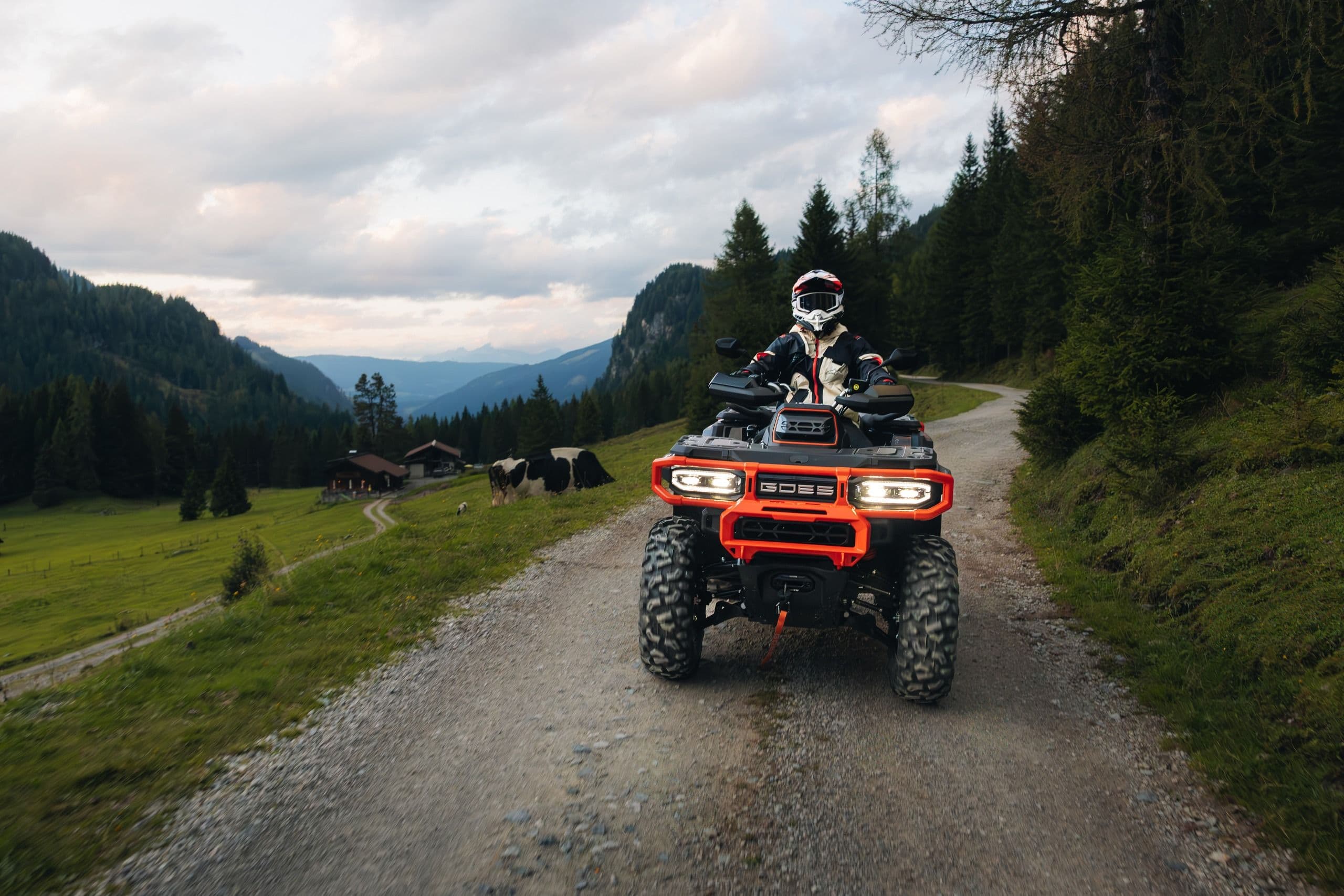 Rider on an orange ATV drives down a dirt road in a green mountain valley.