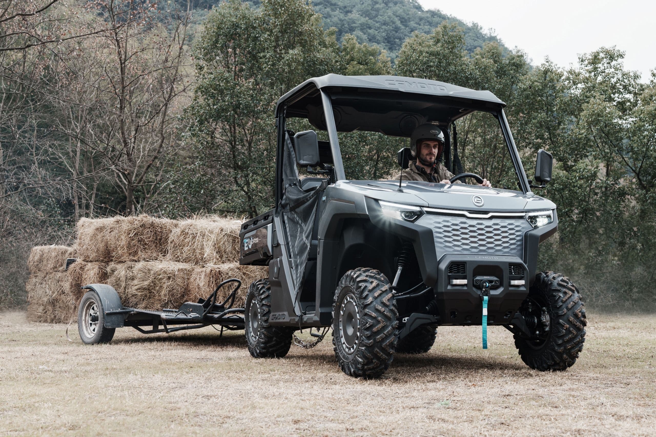 Gray UTV with driver pulls a trailer full of hay bales through a rural field.