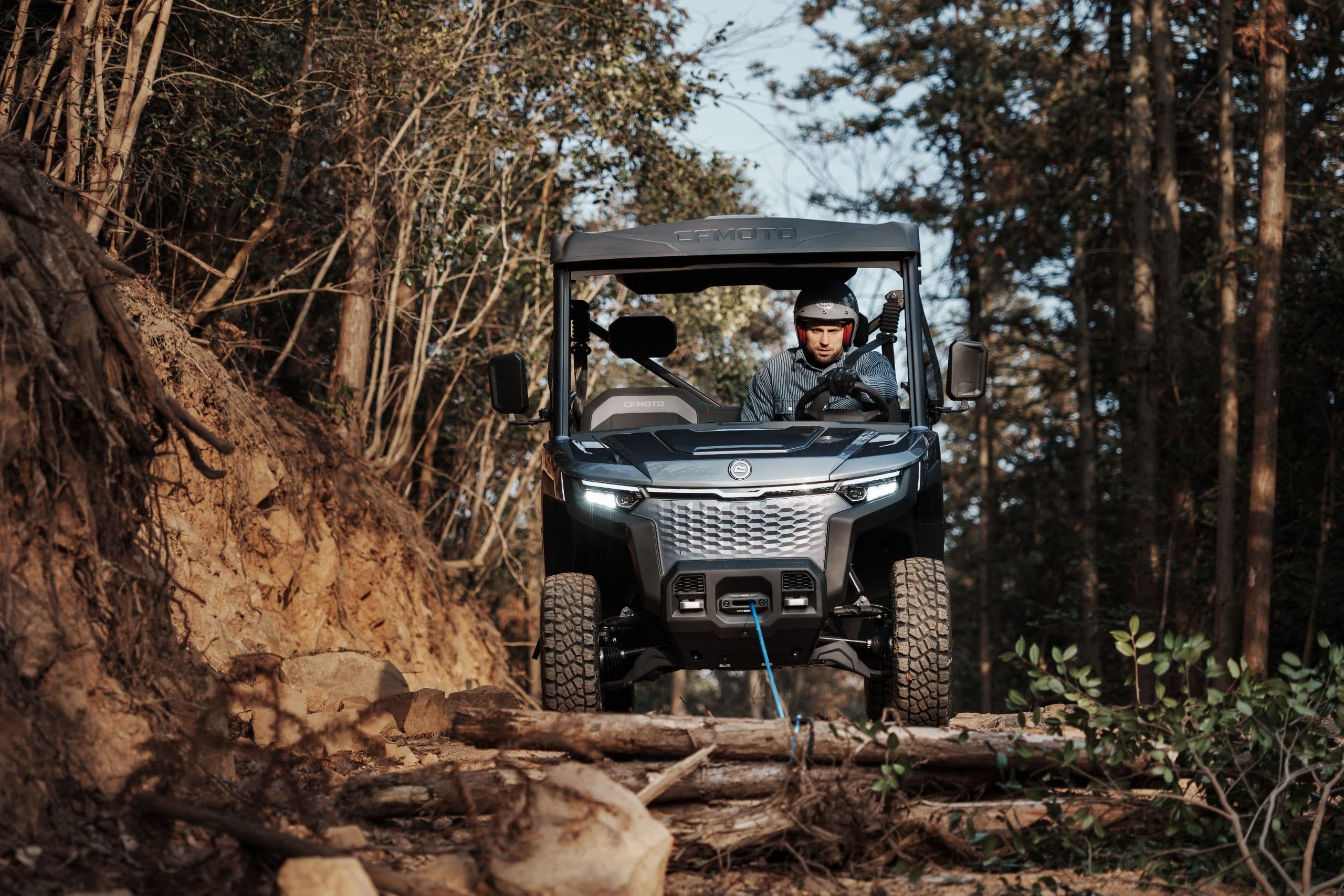 Gray UTV drives on a challenging forested dirt path with logs.