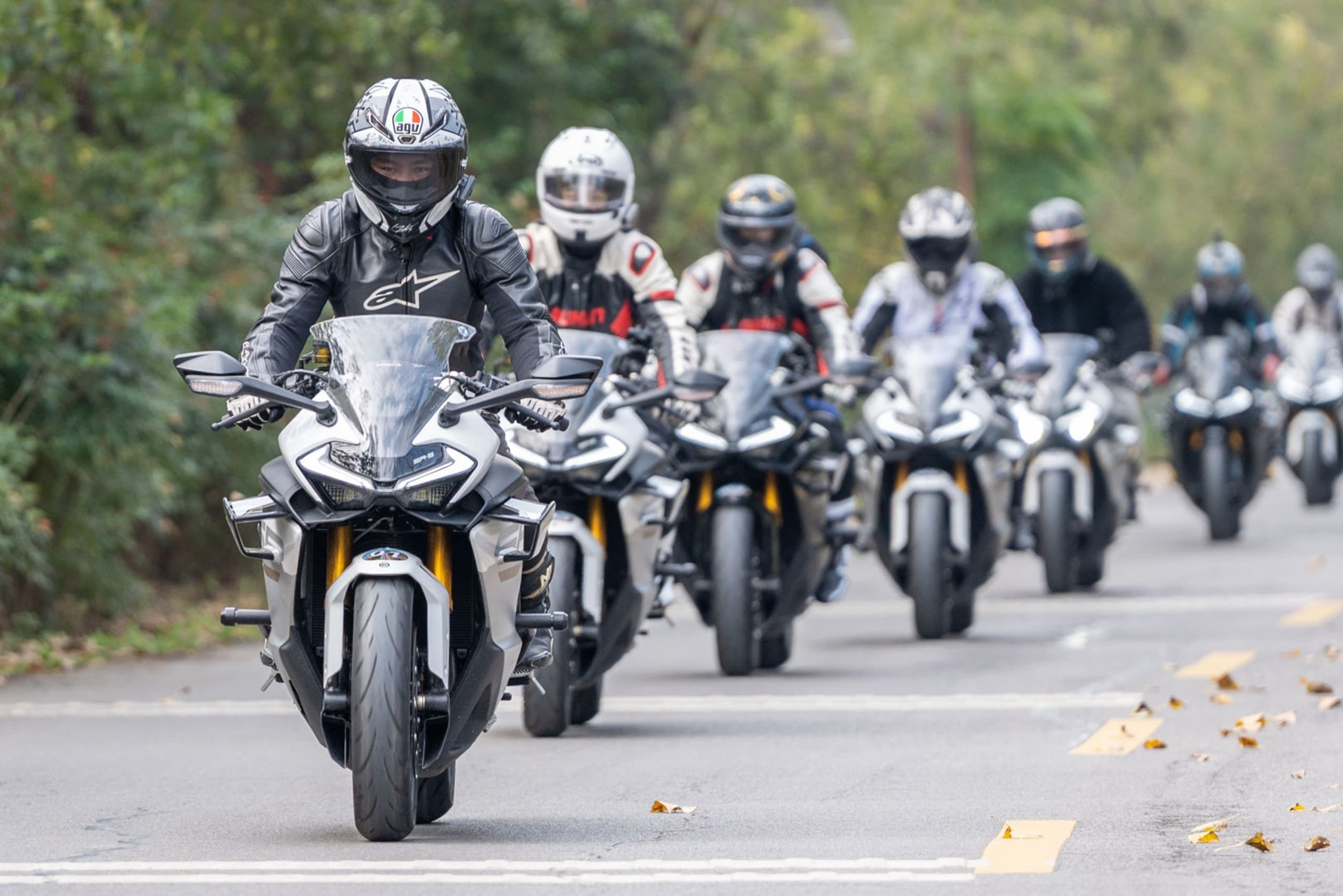 Group of motorcyclists in full gear riding in formation on a tree-lined road.
